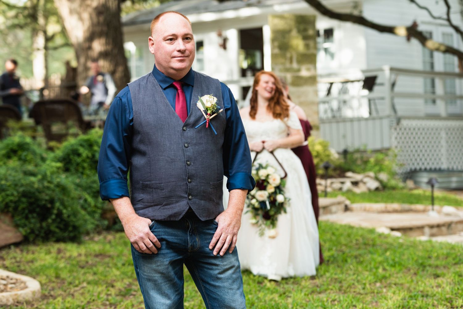 A groom in casual attire with vest and boutonniere stands in a garden setting.