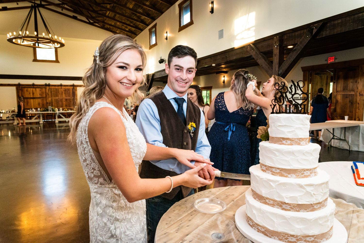 A wedding couple cutting their elegant white tiered cake at their rustic venue reception.