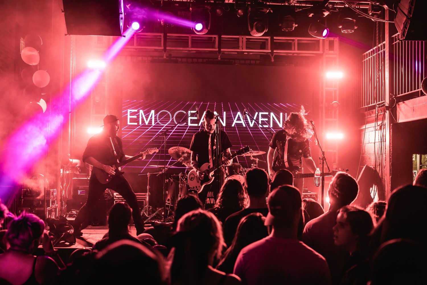 Rock band performs on stage under red and purple lighting with illuminated Emocean Avenue sign.
