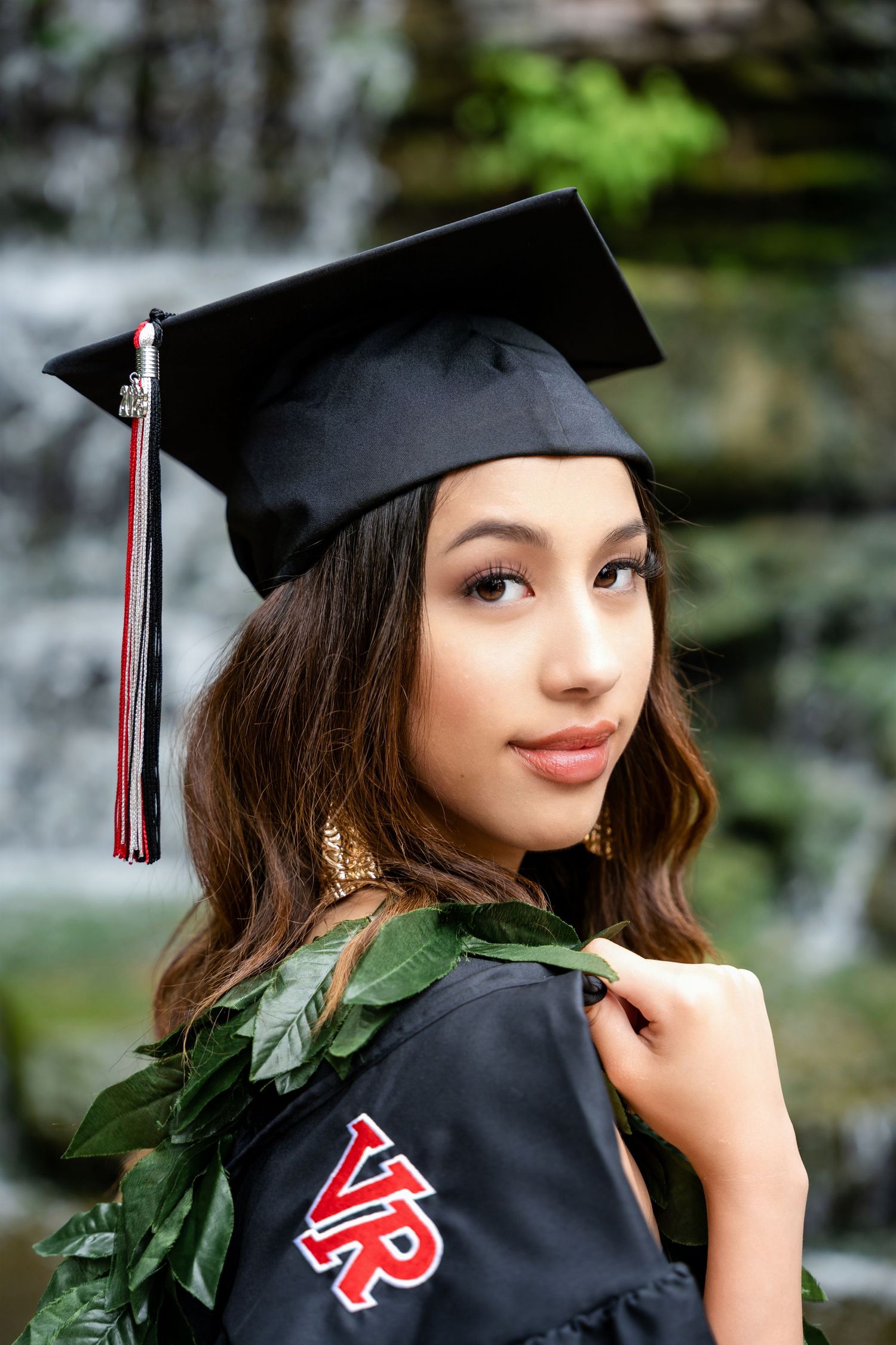 Graduate in cap and gown with VR logo and green lei poses by waterfall.