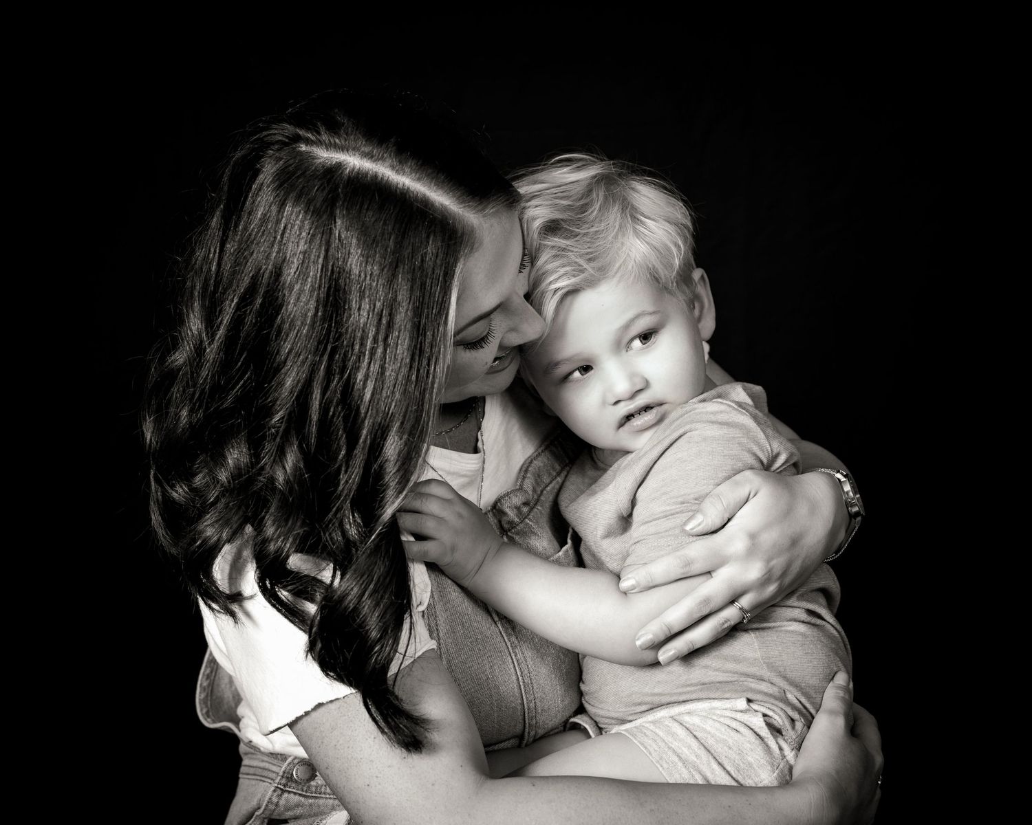 Black and white portrait showing tender embrace between parent and small child against dark background.