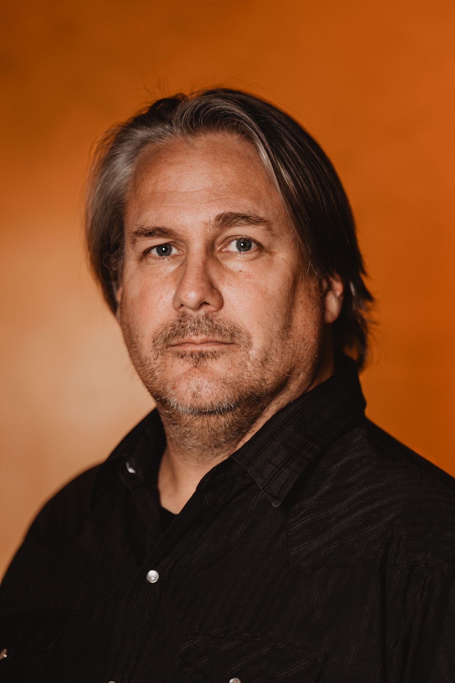 Studio portrait of a man with shoulder-length hair wearing a black shirt against an orange backdrop.