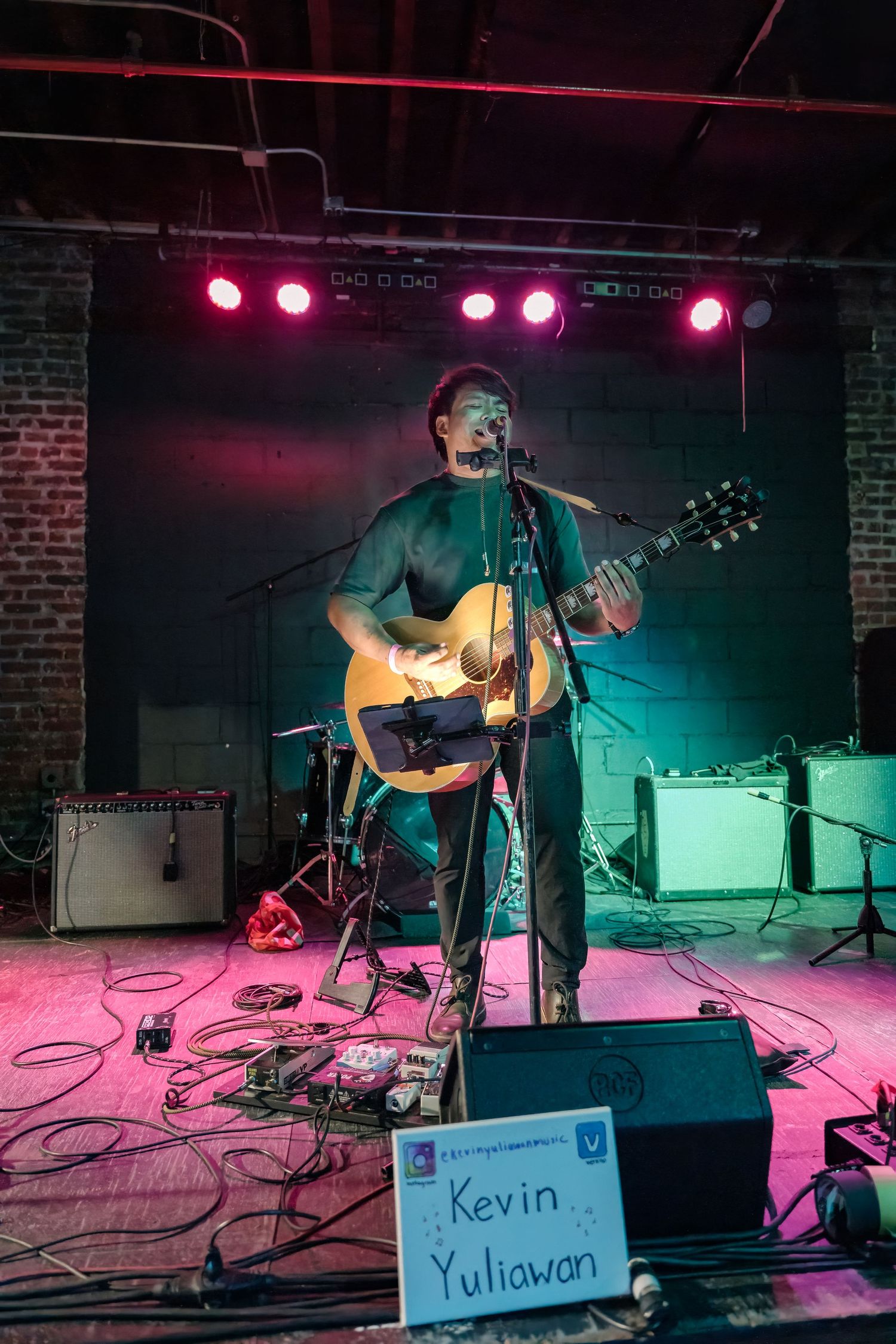 Musician performs on stage with acoustic guitar under red stage lighting with a name sign visible in the foreground.