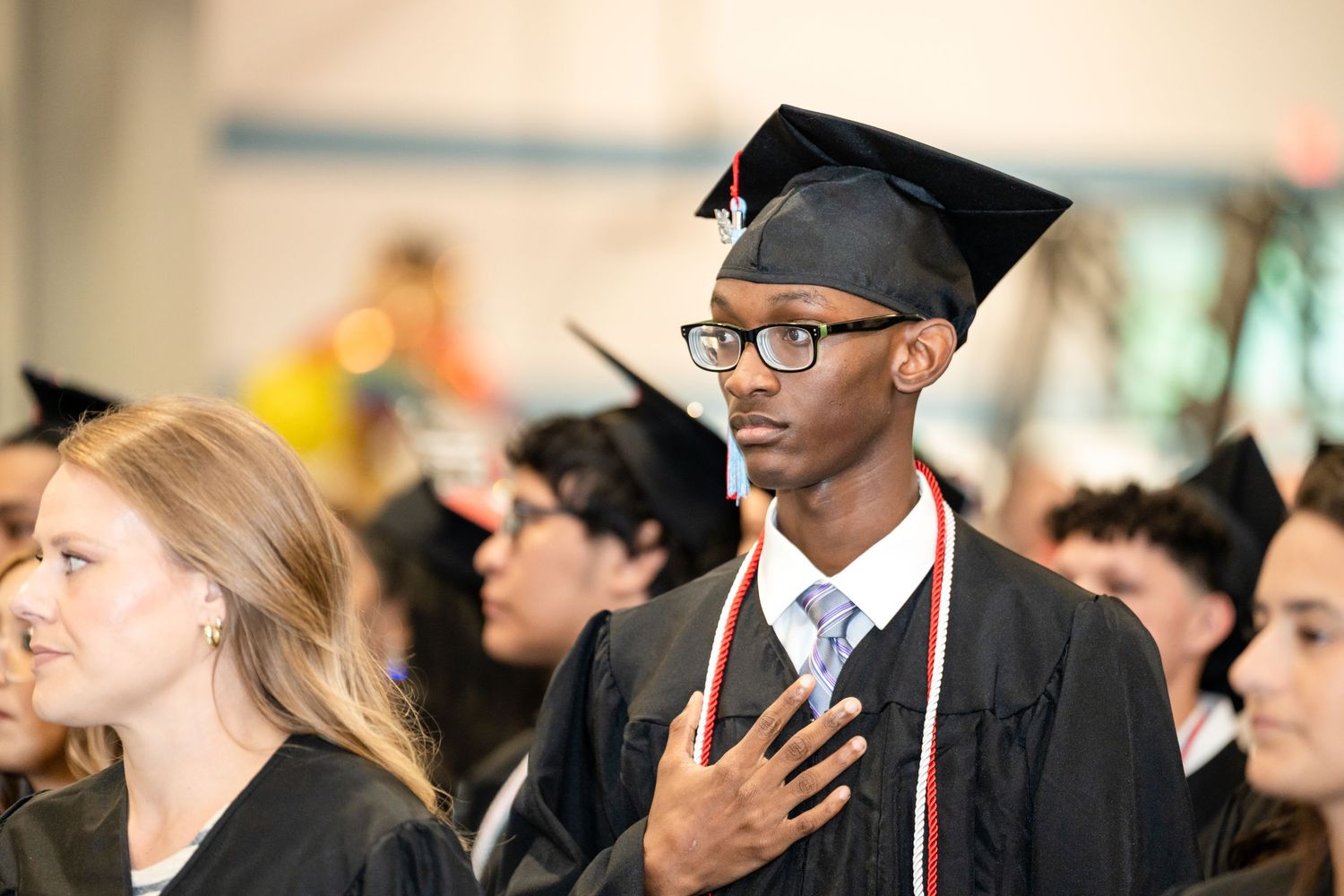 Graduates in academic regalia stand with hands over hearts during commencement.