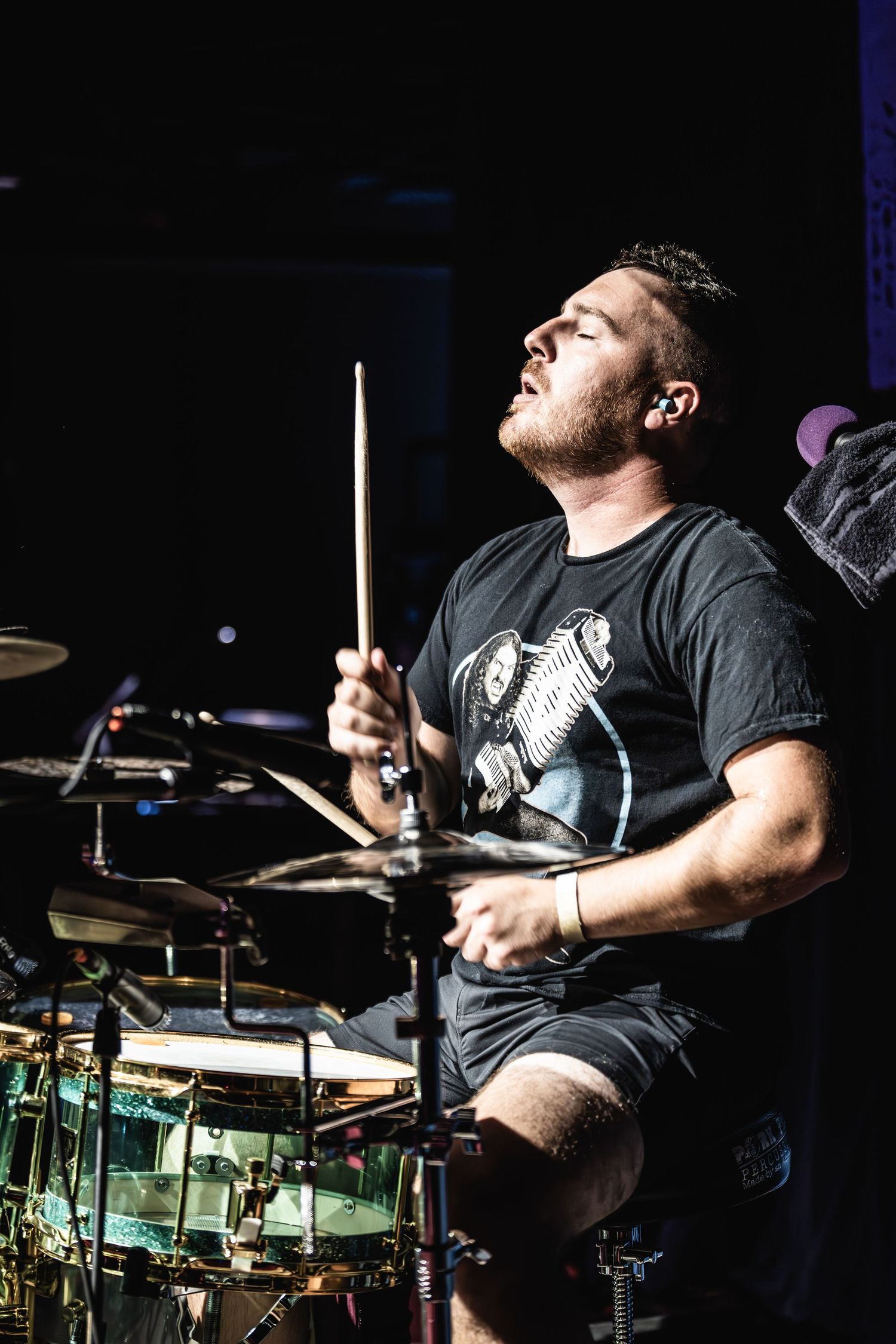 Drummer intensely plays drum kit on stage in dramatic lighting.