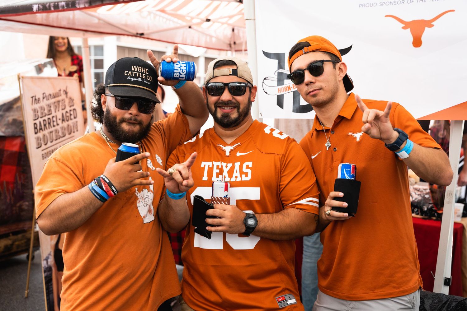 Three Texas Longhorns fans in orange jerseys hold up beverages at a tailgate party.