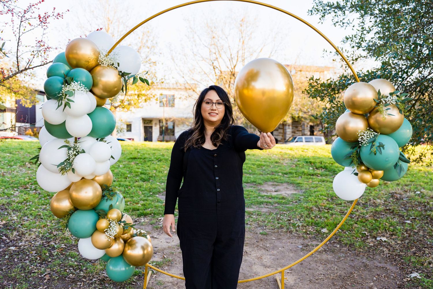 Person standing beneath a circular gold balloon arch with mint and white accents in a garden setting.