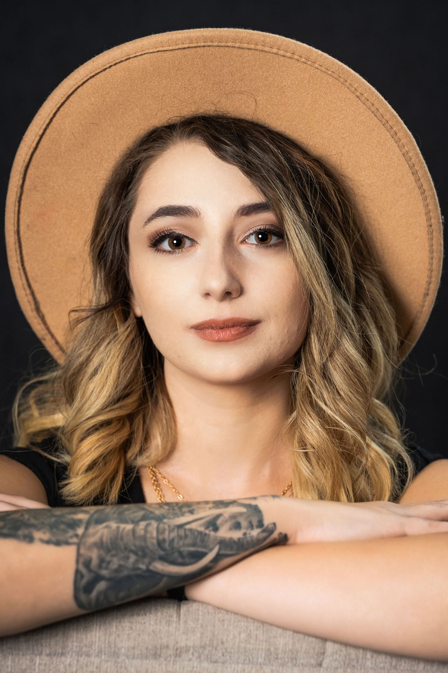 Close-up portrait wearing tan fedora with wavy hair and tattooed arm visible.
