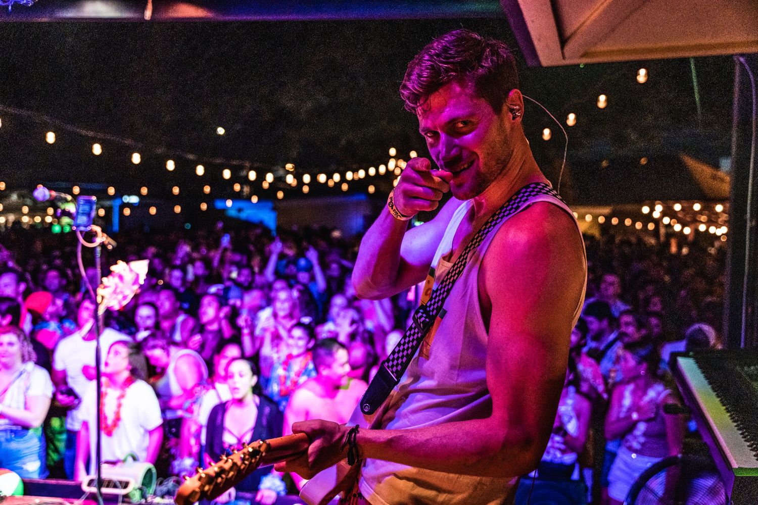 Performer bathed in pink lighting plays on stage with string lights and crowd in background at outdoor concert venue.