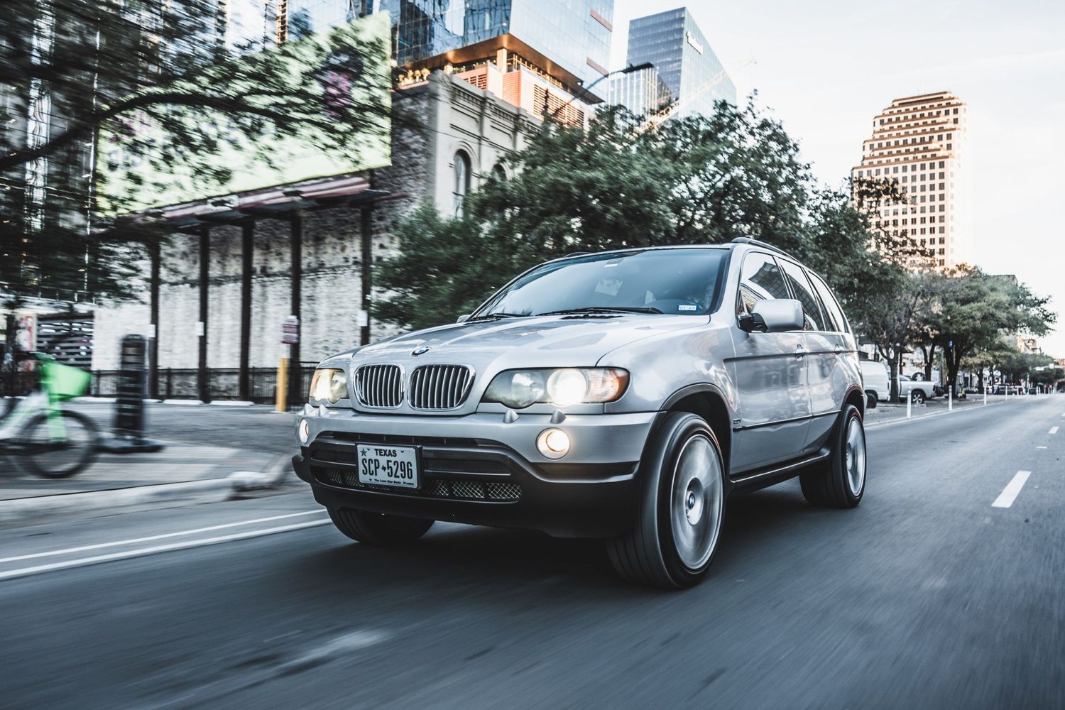 BMW X5 SUV driving through urban street with trees and buildings along sidewalk.