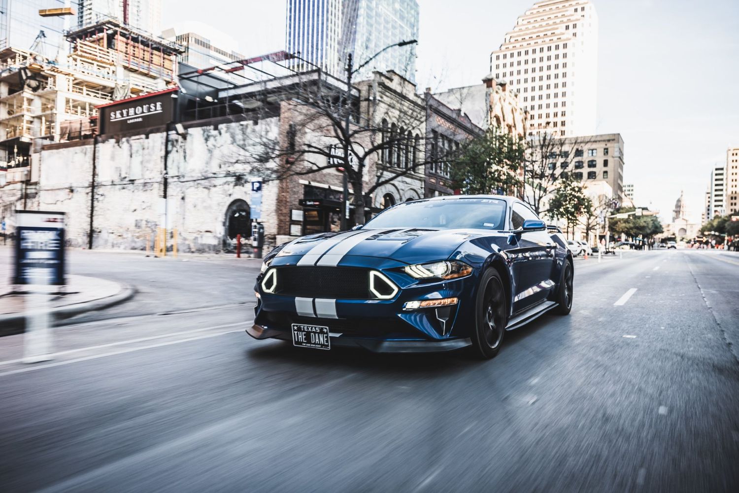 Blue Ford Mustang GT with performance modifications speeds through downtown urban setting.