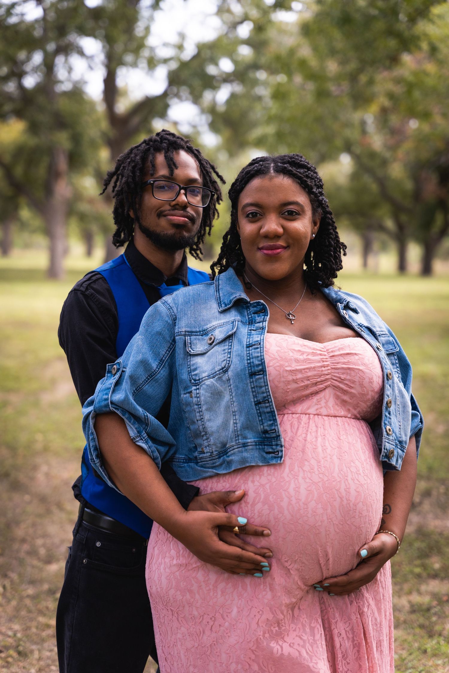 A romantic maternity photoshoot in a park featuring a couple in casual attire with a blue denim jacket and pink dress.