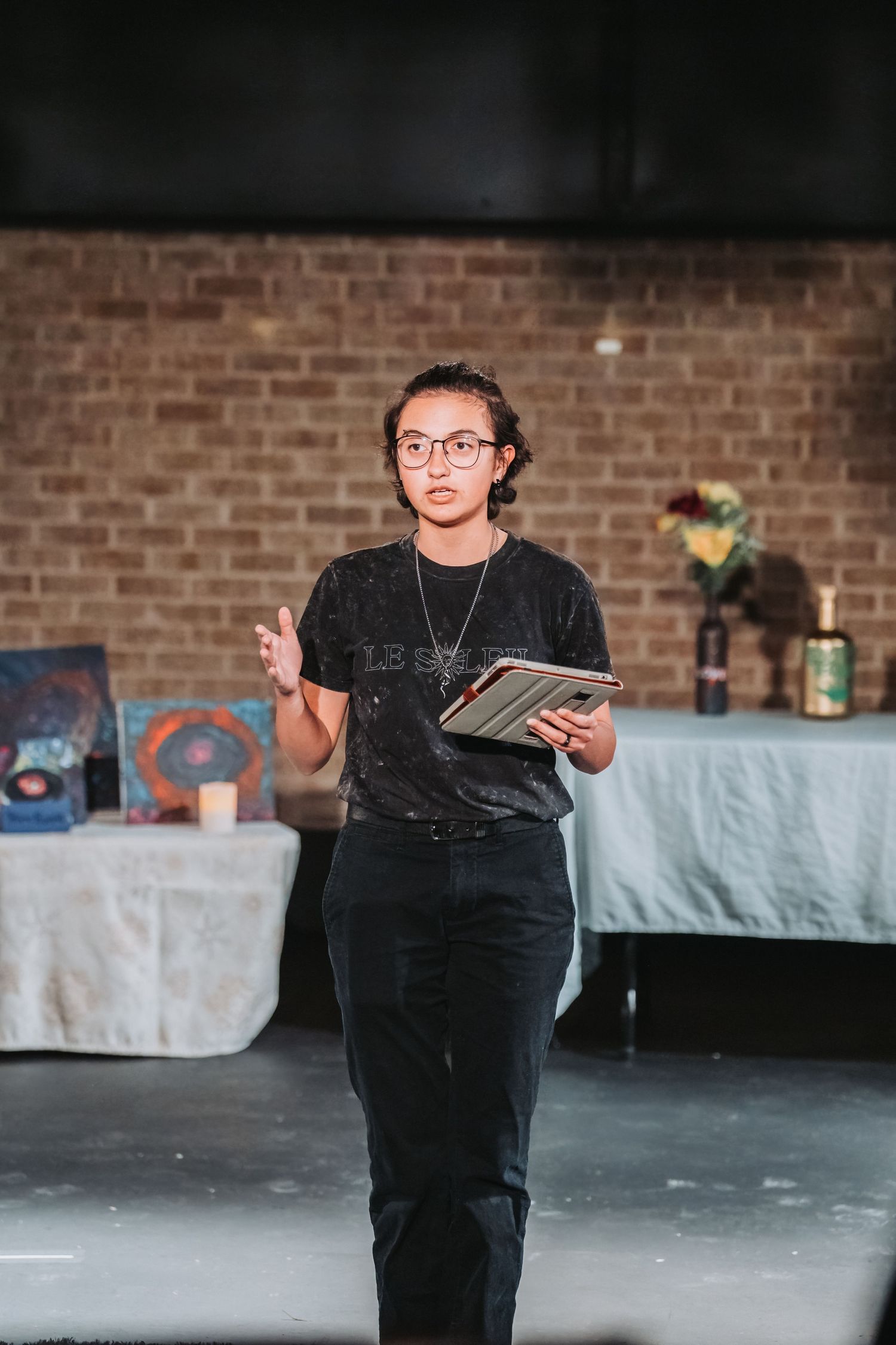 A performer in black attire holds a book while speaking on stage with a brick wall backdrop.