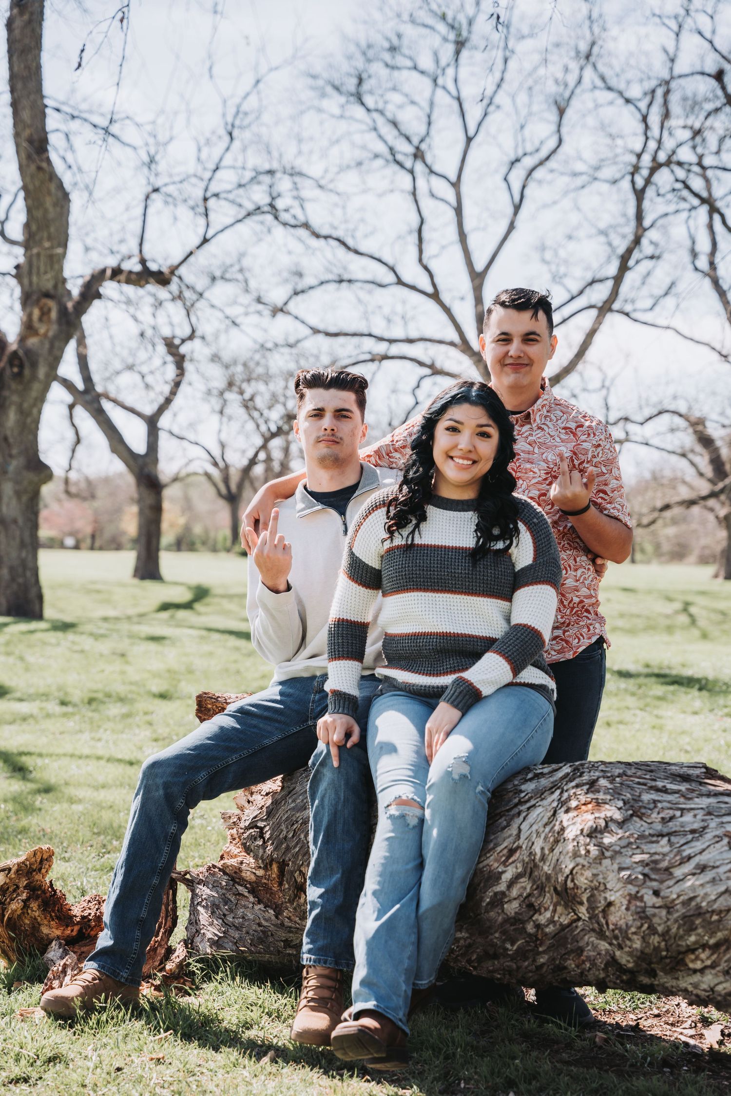 Three people sit together on fallen tree trunk in park setting with bare trees.