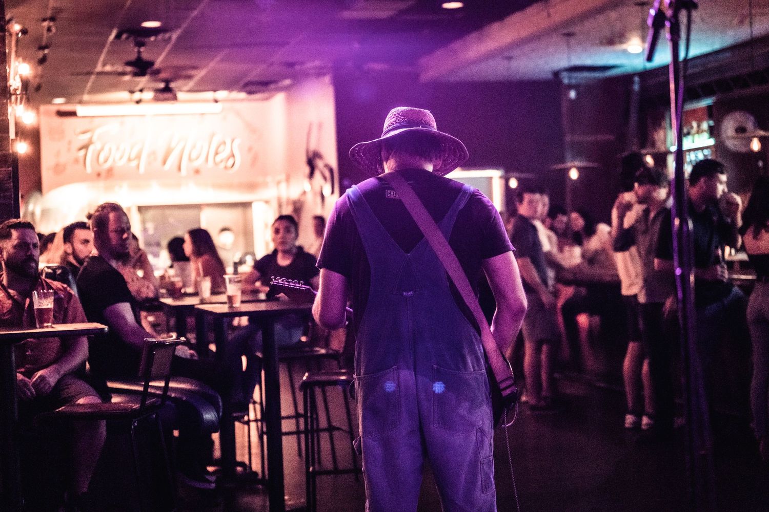 Musician wearing a cowboy hat performing at a dimly lit bar venue with patrons seated at tables.