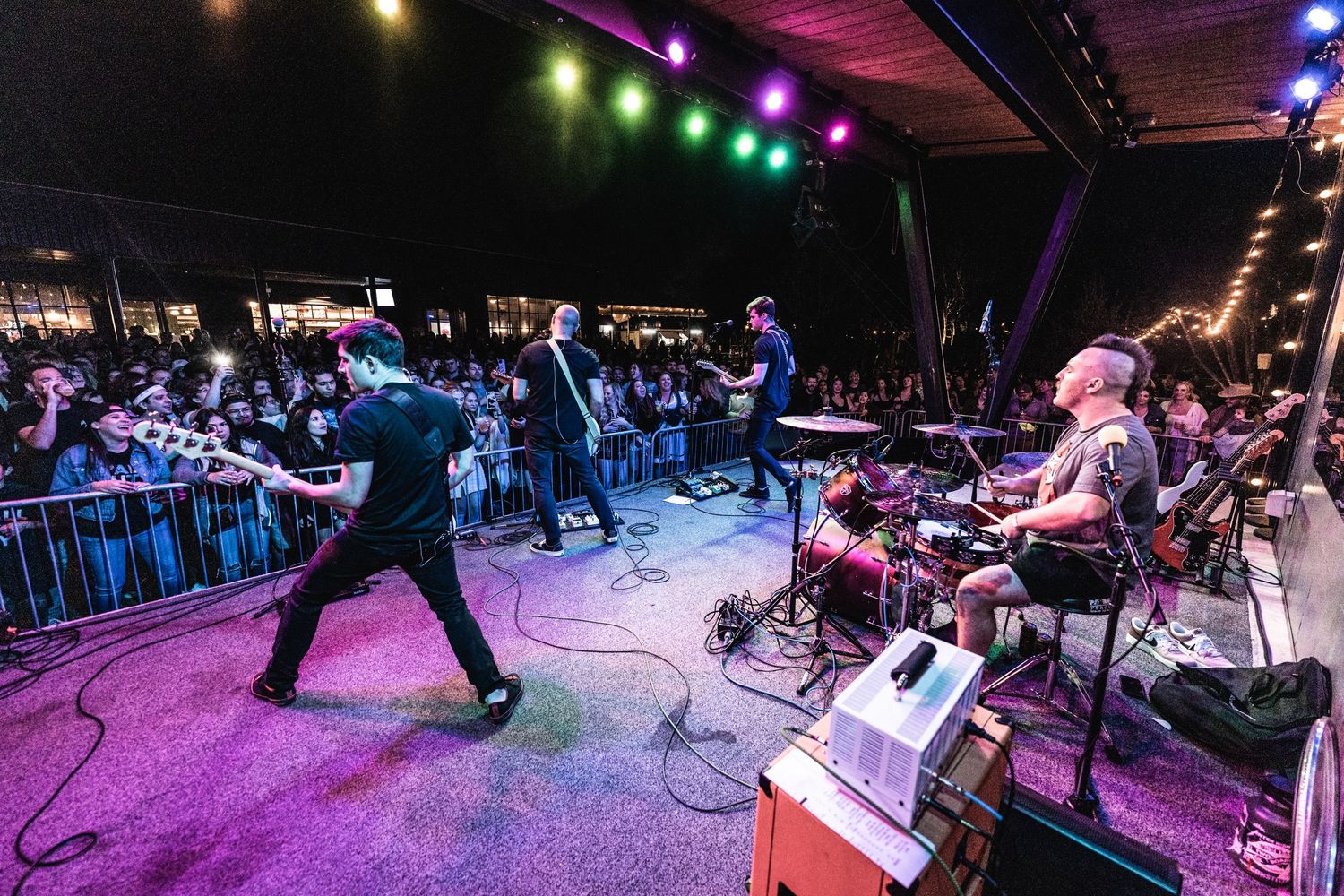 Band performs on outdoor stage with colorful lighting as crowd watches behind security barriers.
