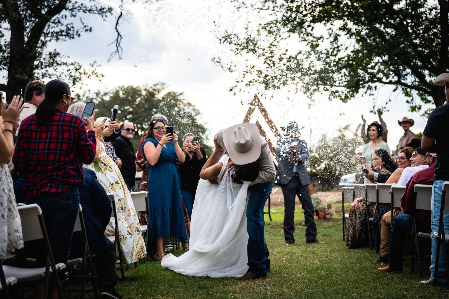 Wedding guests celebrate as newlyweds walk down the aisle at outdoor ceremony.