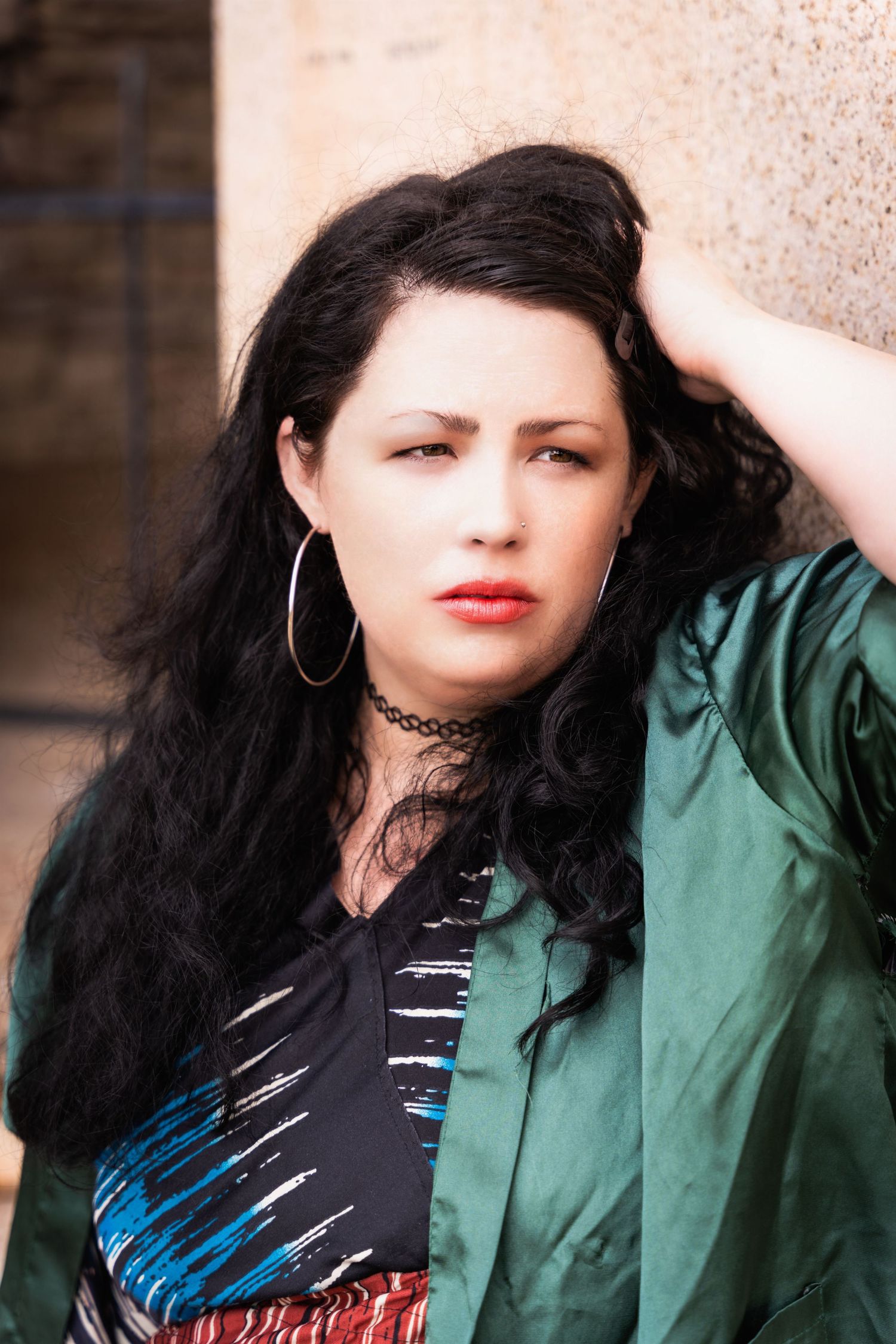 Close-up portrait with long dark hair wearing green jacket and choker against stone wall background.