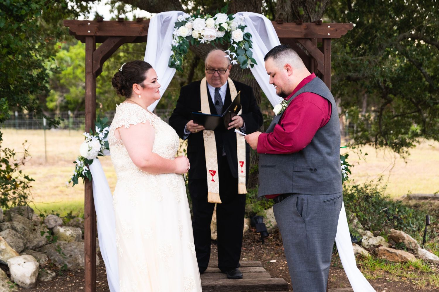Outdoor wedding ceremony taking place under wooden arch decorated with white fabric and floral garlands.