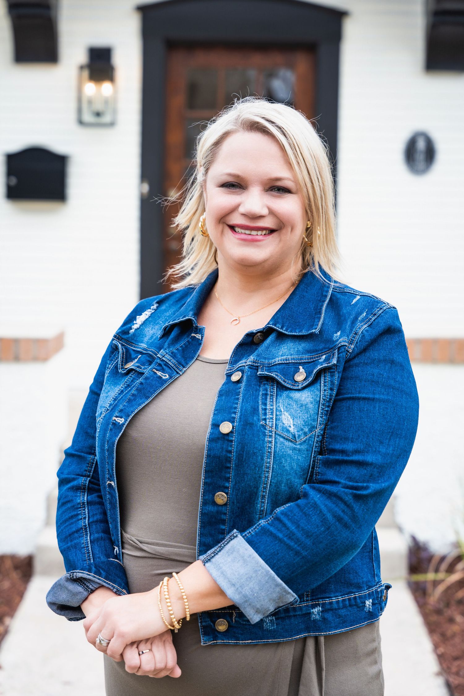Person in denim jacket and gray top smiling against white building with black door.