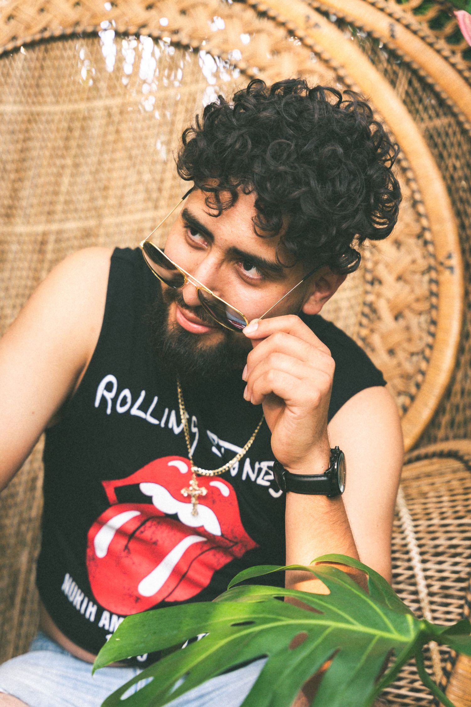Person in Rolling Stones tank top with curly hair relaxes in wicker peacock chair with sunglasses.
