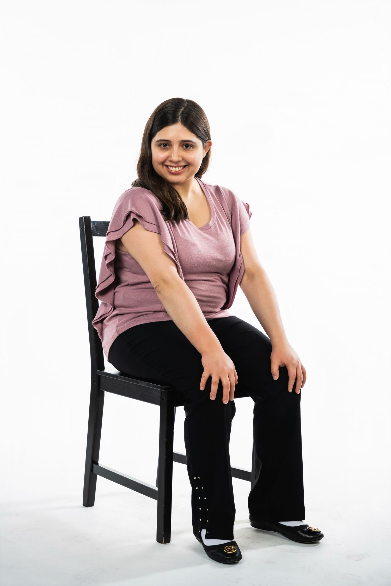 Studio portrait of someone in a mauve top sitting on a black chair against white background.