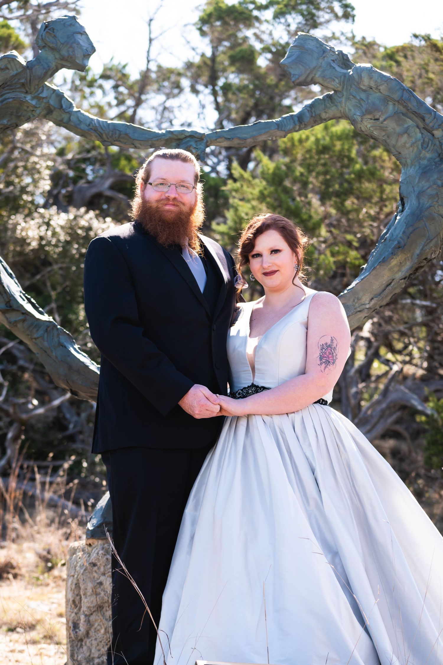 Couple in formal wedding attire pose under a unique metal arch sculpture outdoors.