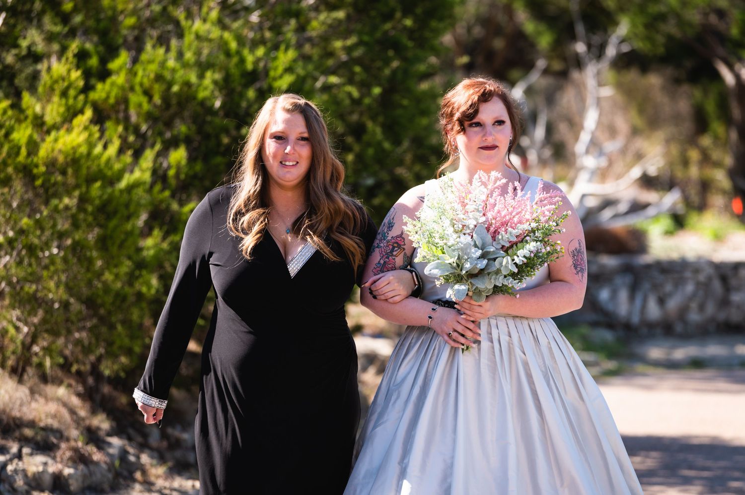 Two people walk outdoors during a wedding, one wearing a light-colored bridal gown.