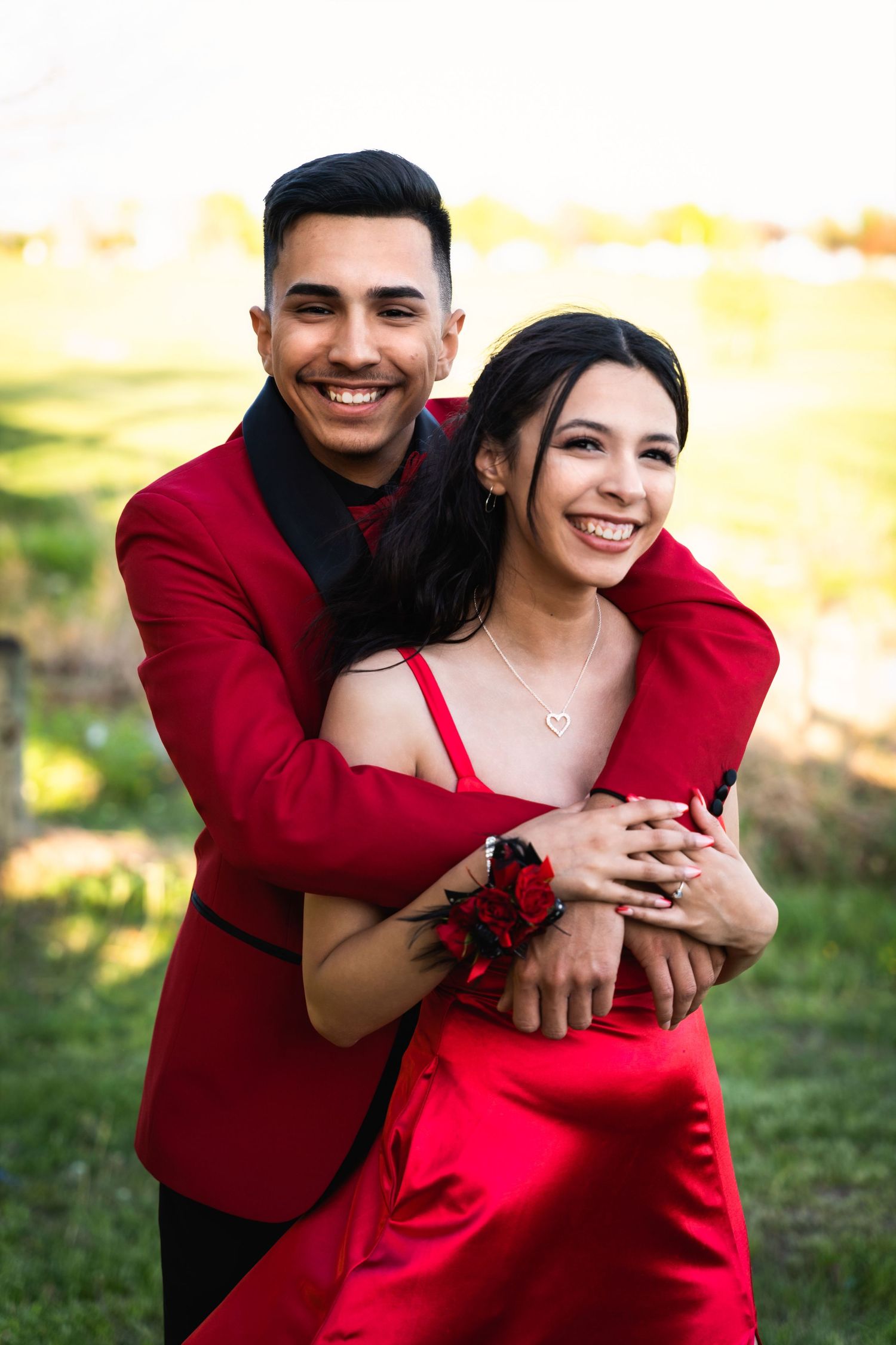 Couple in matching red formal wear embrace outdoors in sunny garden setting.