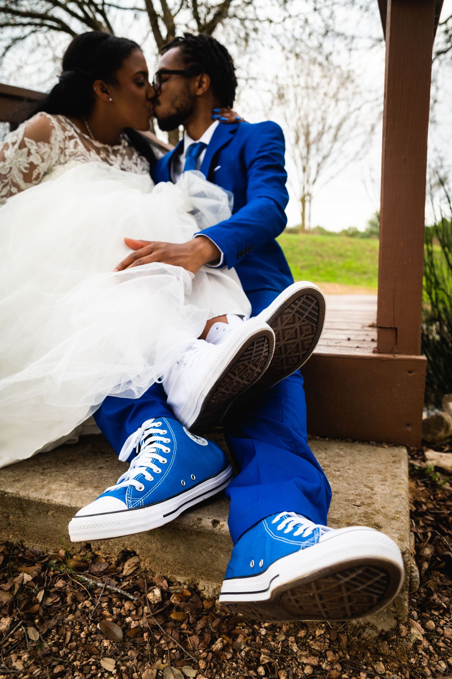 Wedding couple wearing matching blue Converse sneakers with formal attire on wooden deck.