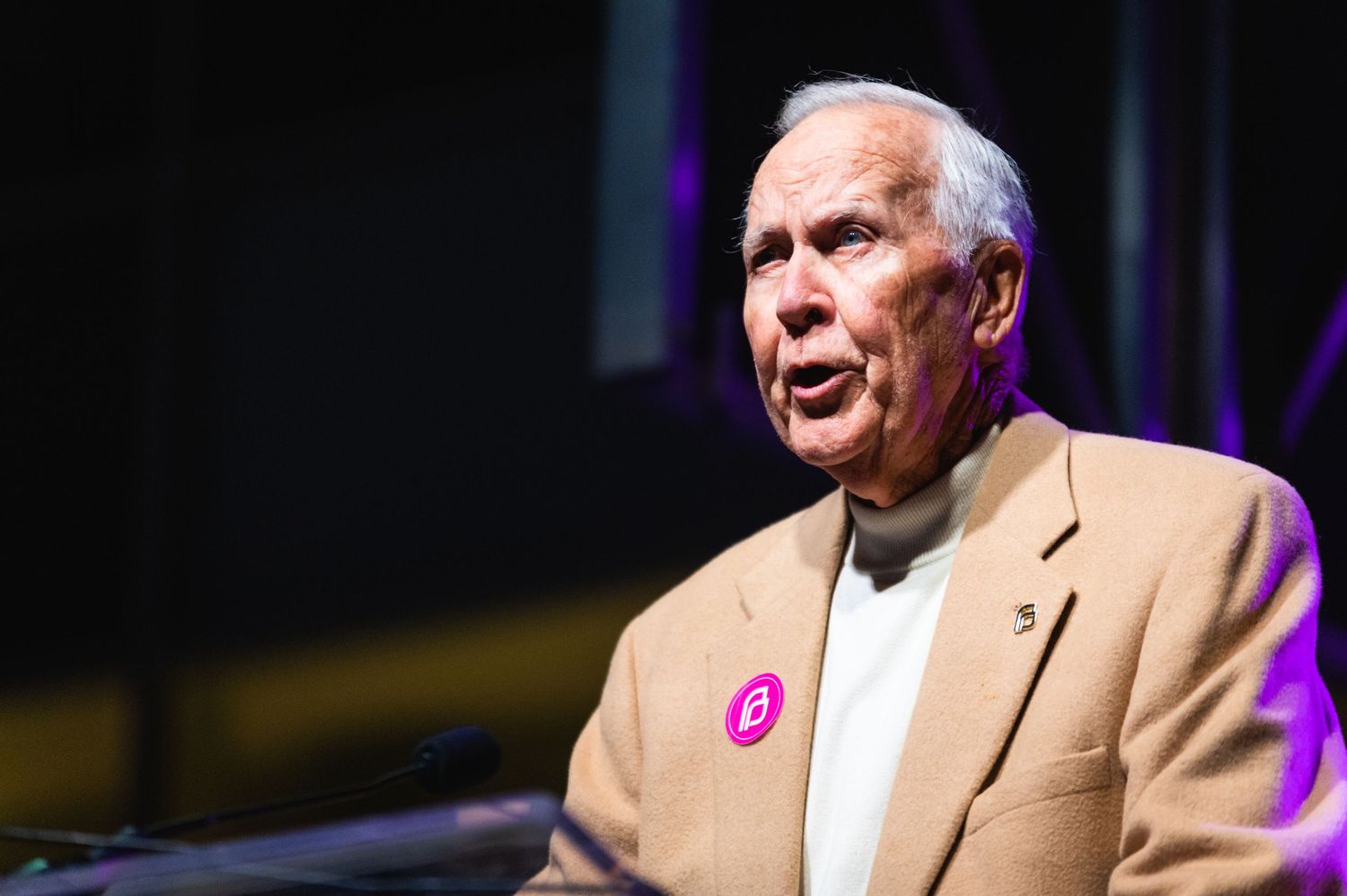 Speaker in tan jacket with pink pin addresses audience from podium under dramatic lighting.