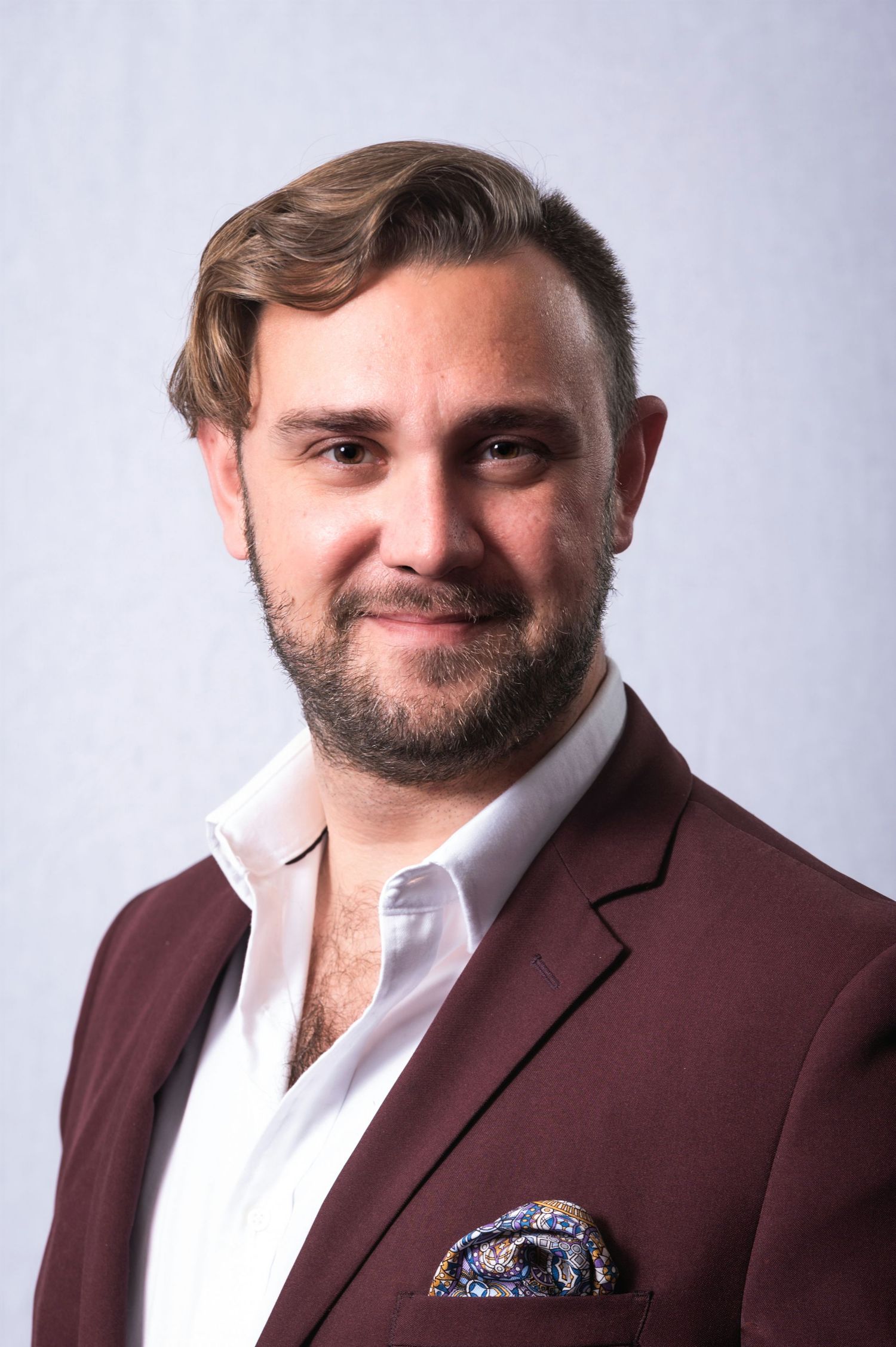 Professional headshot of someone wearing a burgundy blazer and white shirt against a gray background.