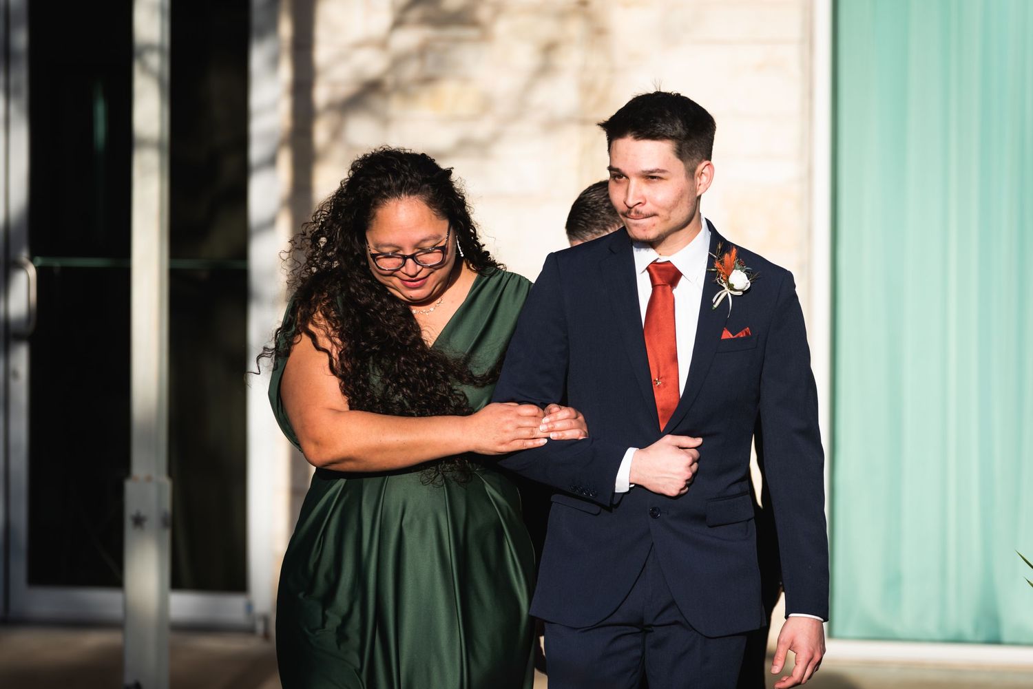 Wedding party members walk arm in arm outside venue in formal attire.