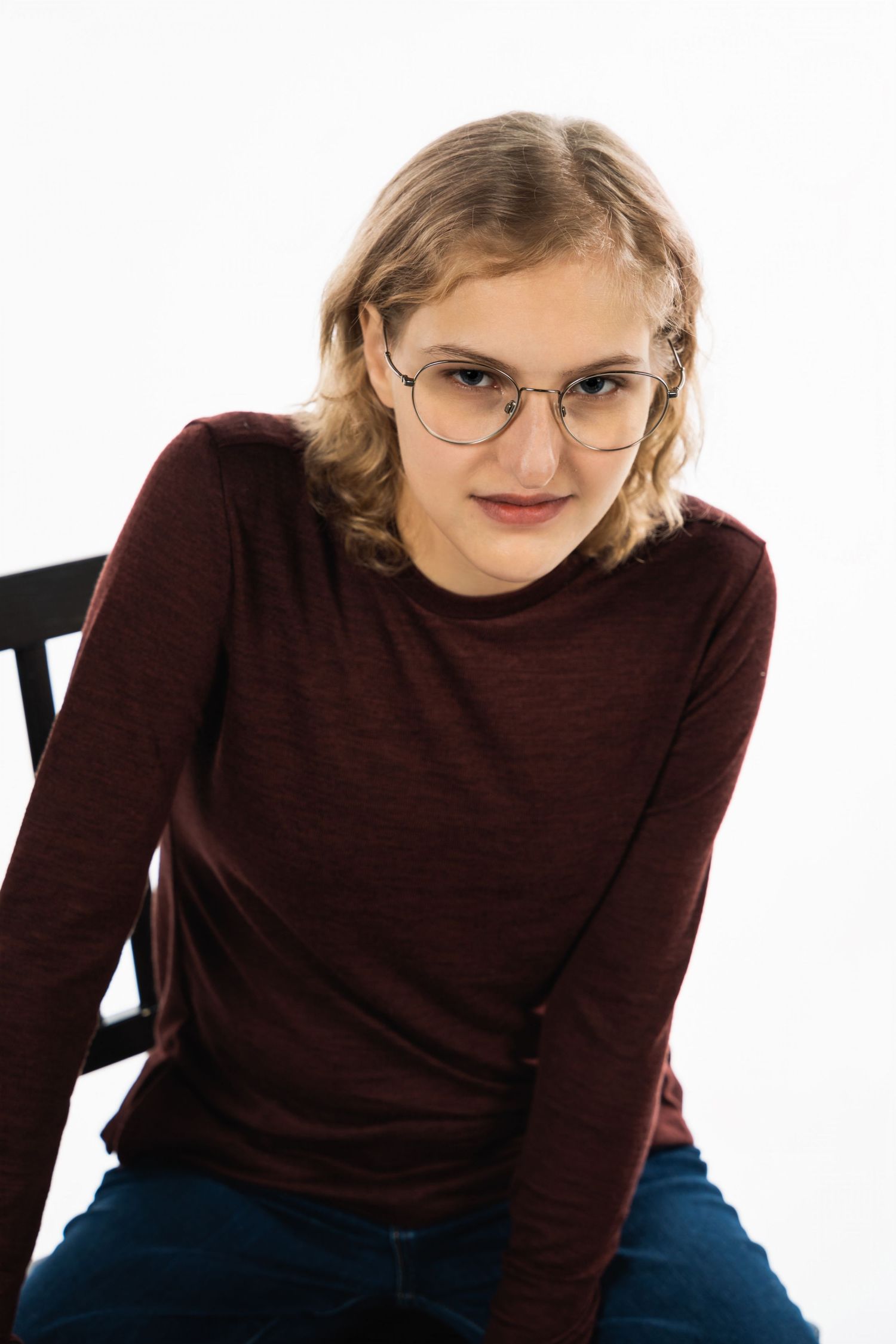 Young woman in burgundy sweater and glasses posing on a chair against white background.