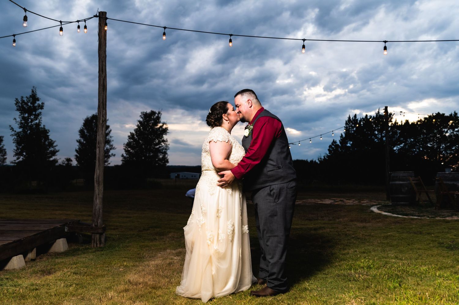 A couple shares a romantic moment under string lights during sunset at an outdoor wedding venue.