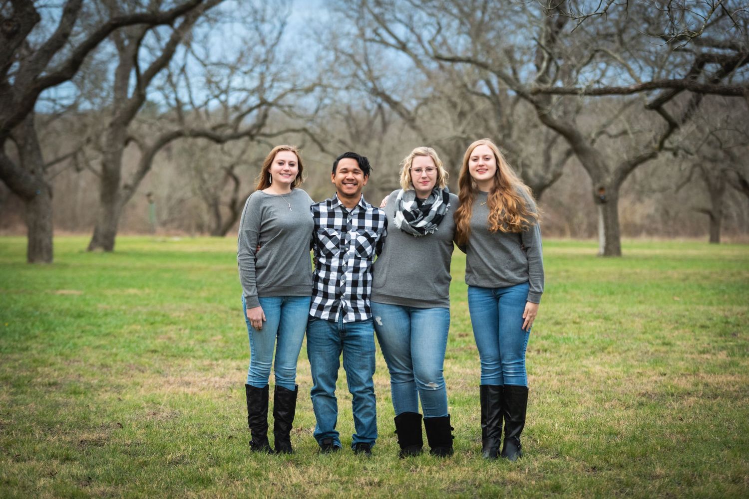Four people in matching jeans and tops pose together in grassy field with trees behind them.