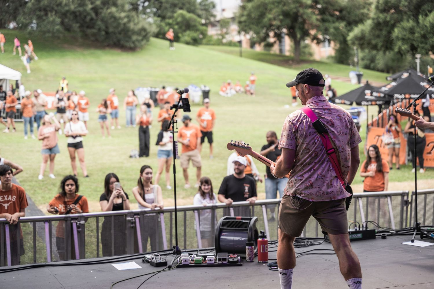 Crowd gathered at an outdoor concert venue with stage equipment and people enjoying live music in a park setting.