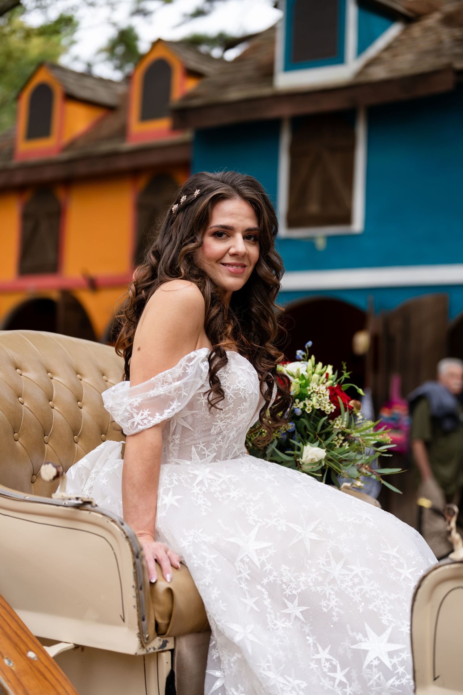 A bride in a white lace wedding dress poses elegantly on a beige tufted chair against colorful buildings.