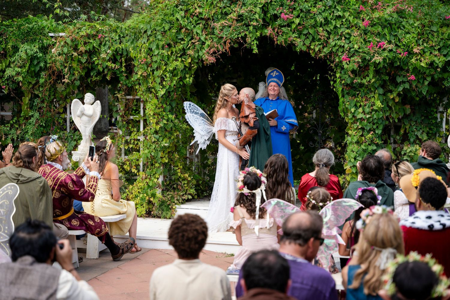 Wedding ceremony takes place in a lush garden setting with guests seated on white chairs and greenery overhead.