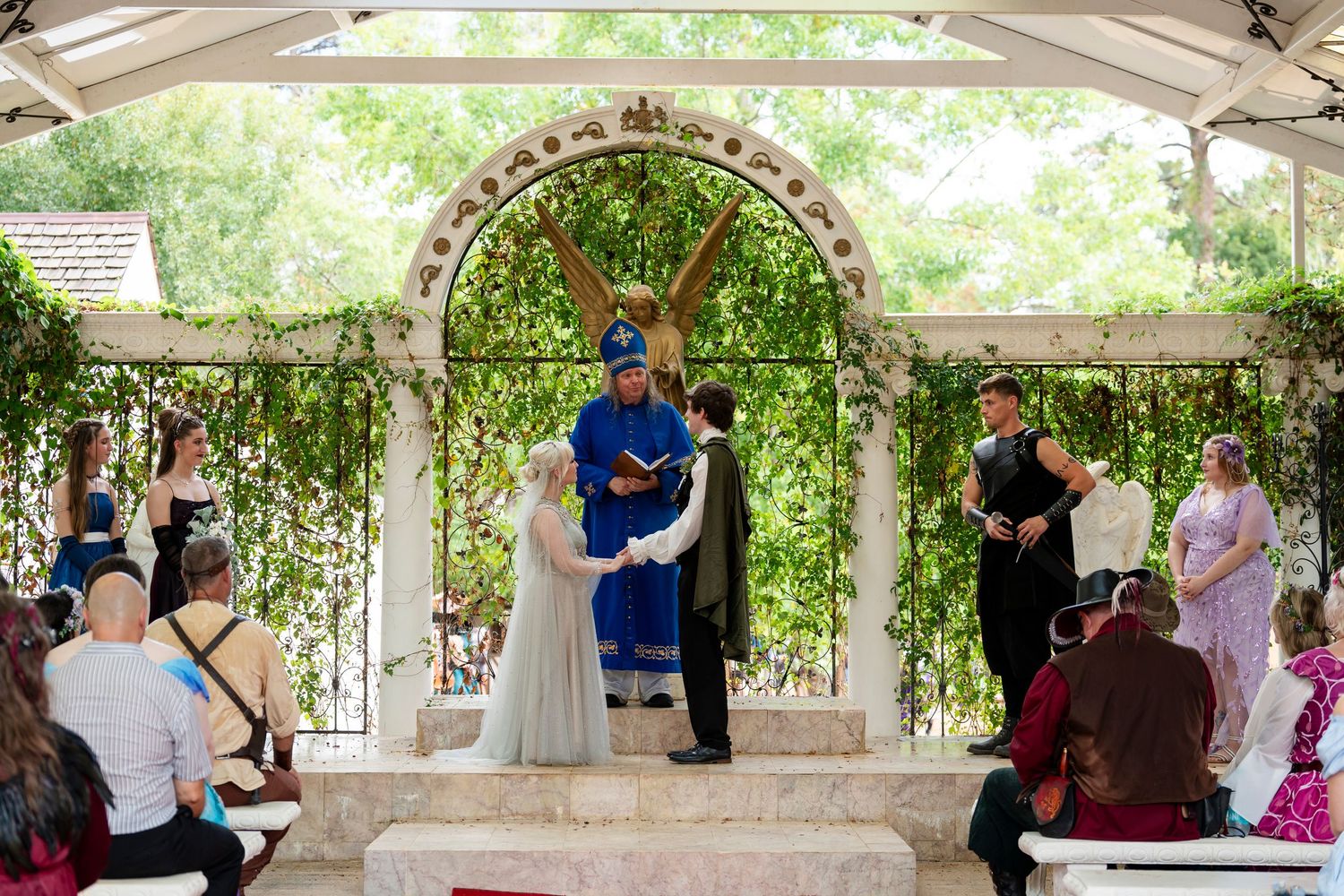 People gather under a white pergola for an outdoor celebration in a garden setting.