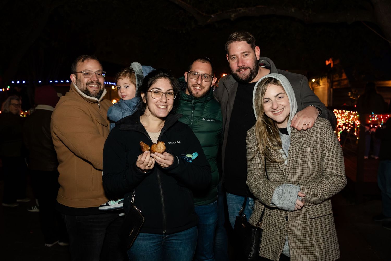 A group of people pose together at a nighttime gathering with festive lights in the background.