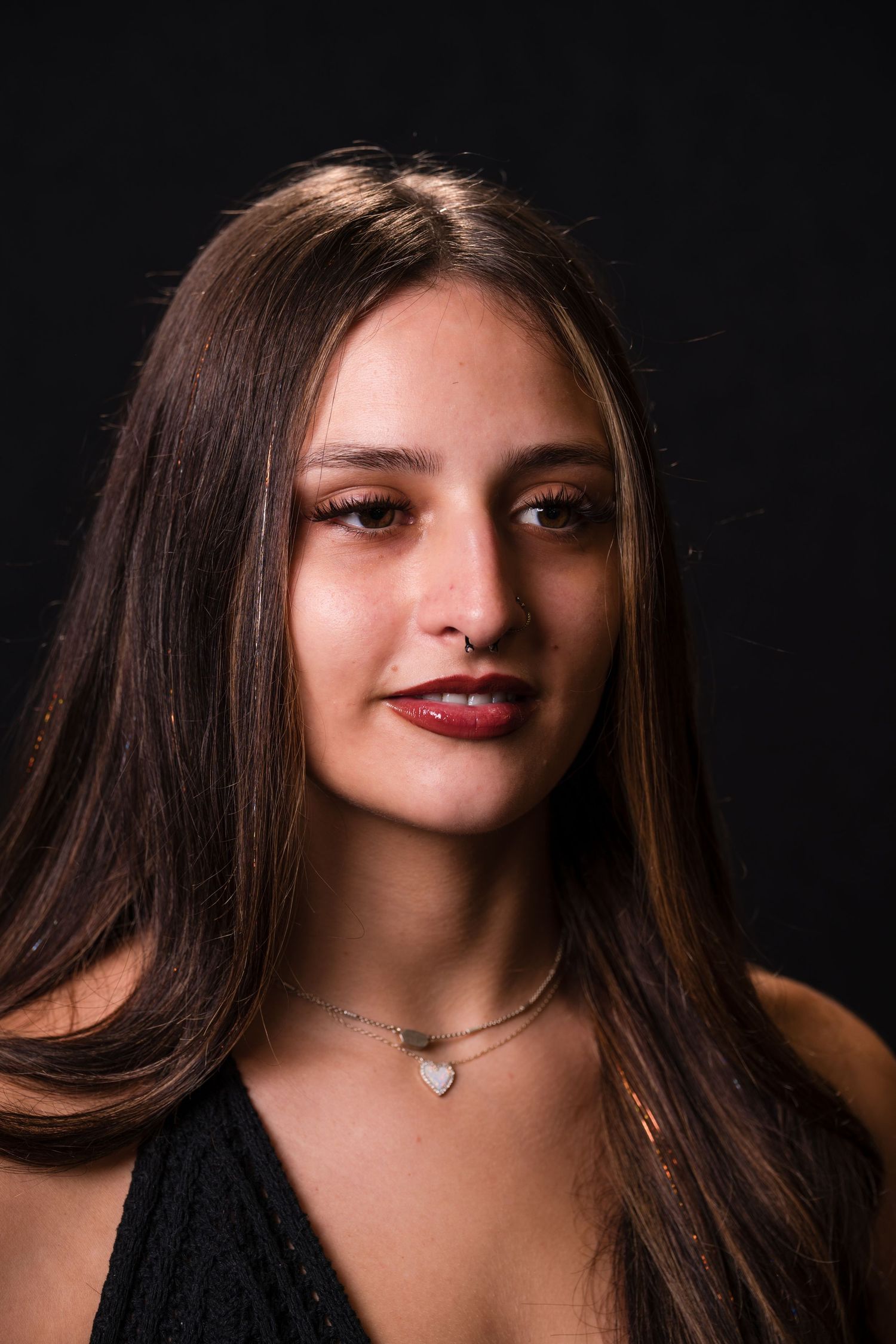 Portrait against dark background showing long brown hair, delicate necklace and red lipstick in dramatic lighting.