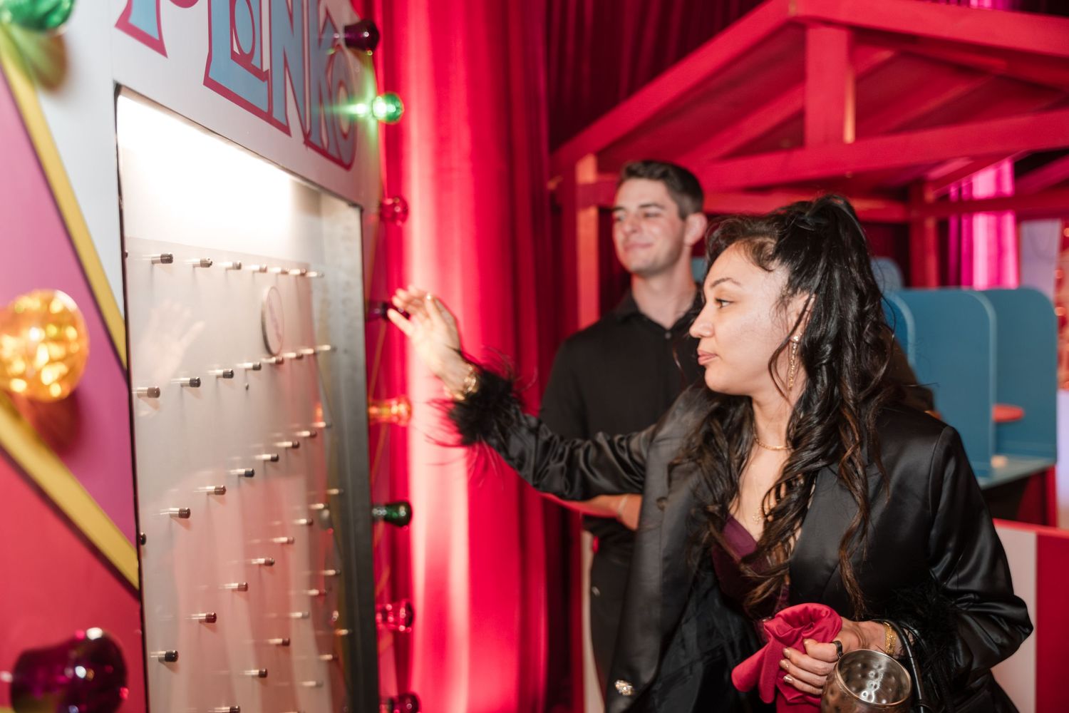 People enjoy drinks and conversation at a vibrant pink-lit bar with neon accents and string lights.