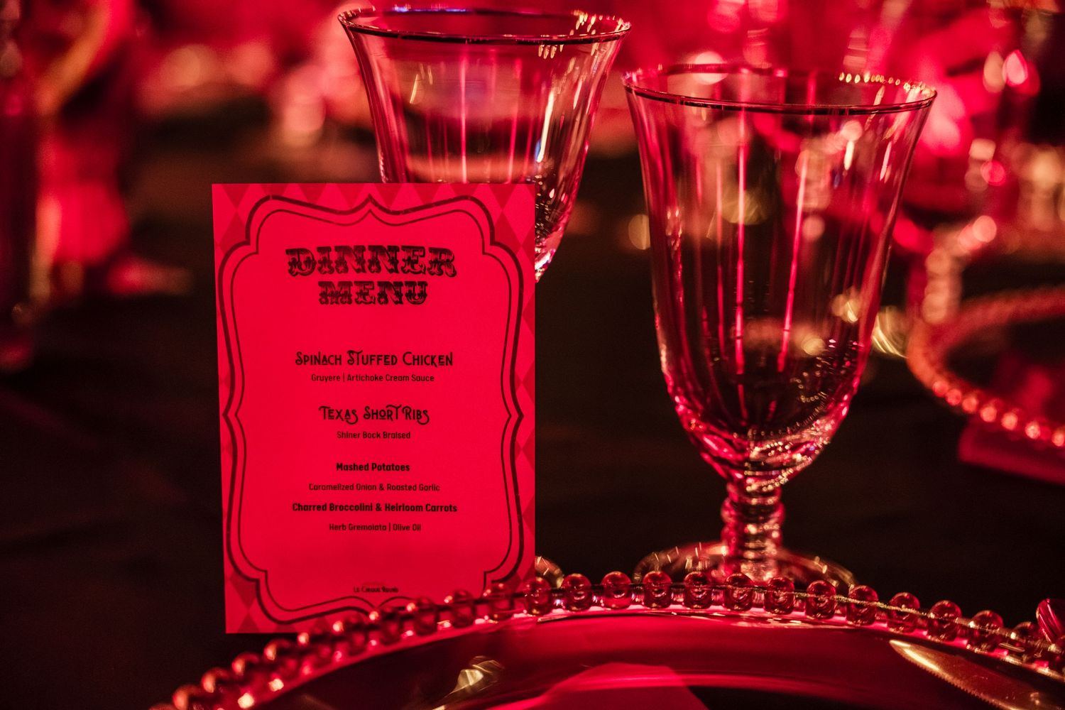 A dinner menu card and elegant wine glass illuminated in red lighting with decorative pearl beads on a dark table.
