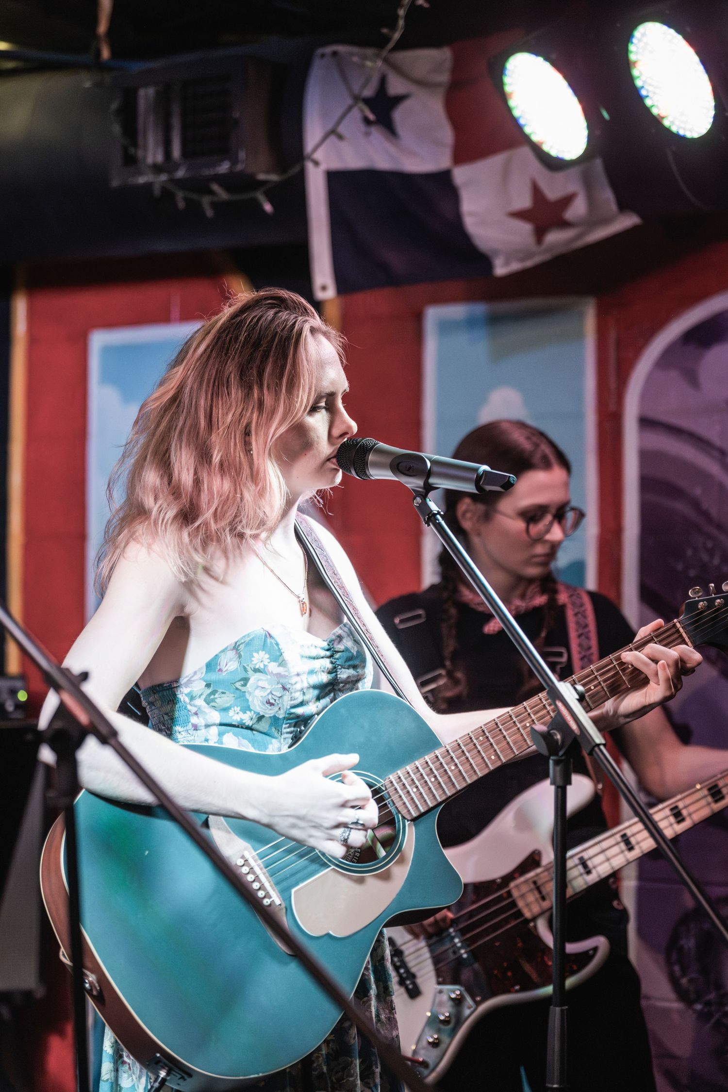 Musician performs on stage with a turquoise acoustic guitar under stage lights with a flag hanging in the background.
