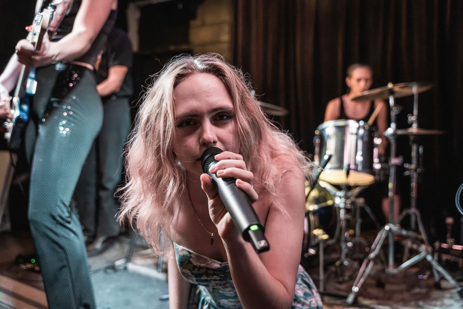 A singer performs passionately on stage with blonde hair and a microphone while a drummer plays in the background.