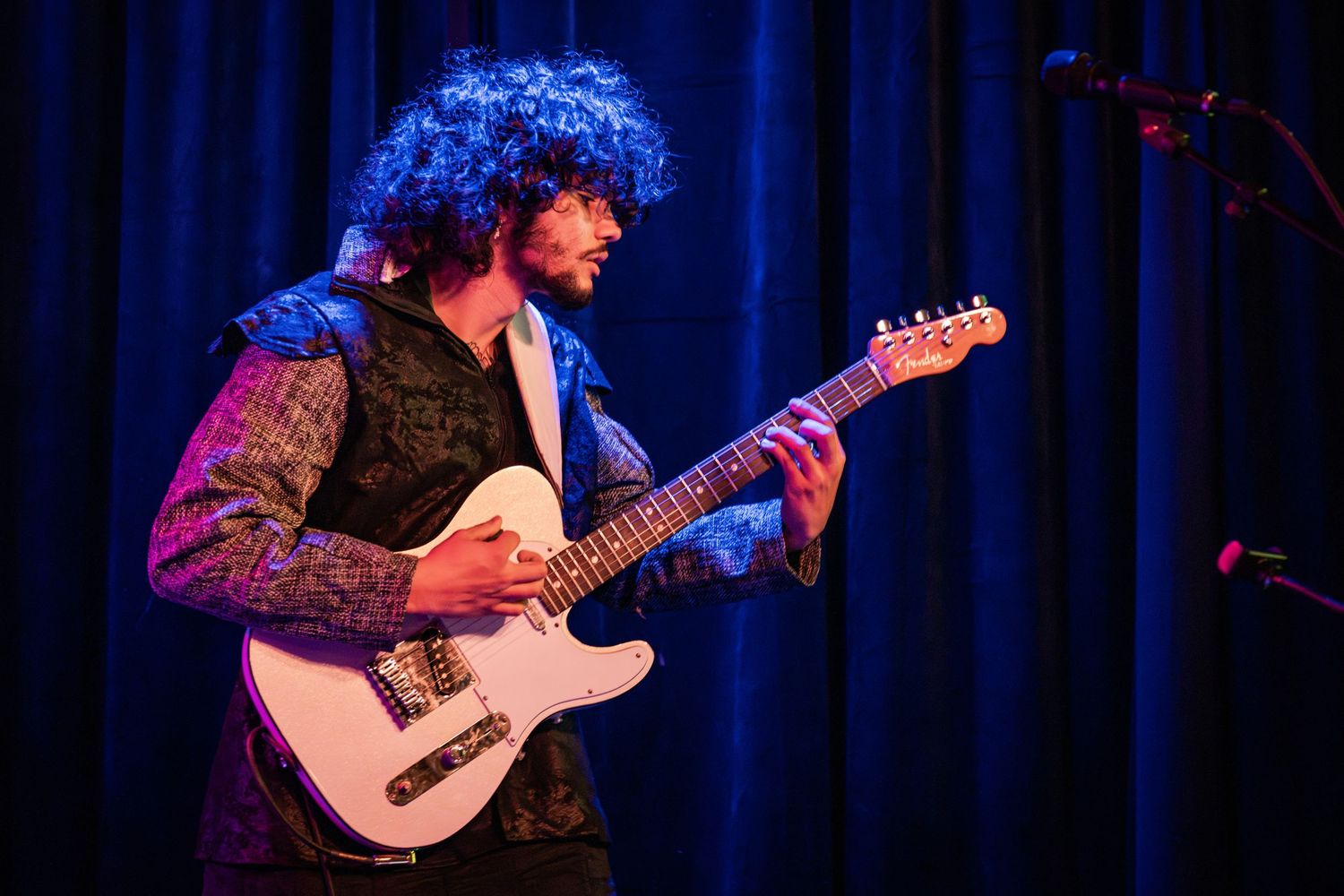 Musicians perform on stage with electric guitars under blue and purple stage lighting during a live concert.