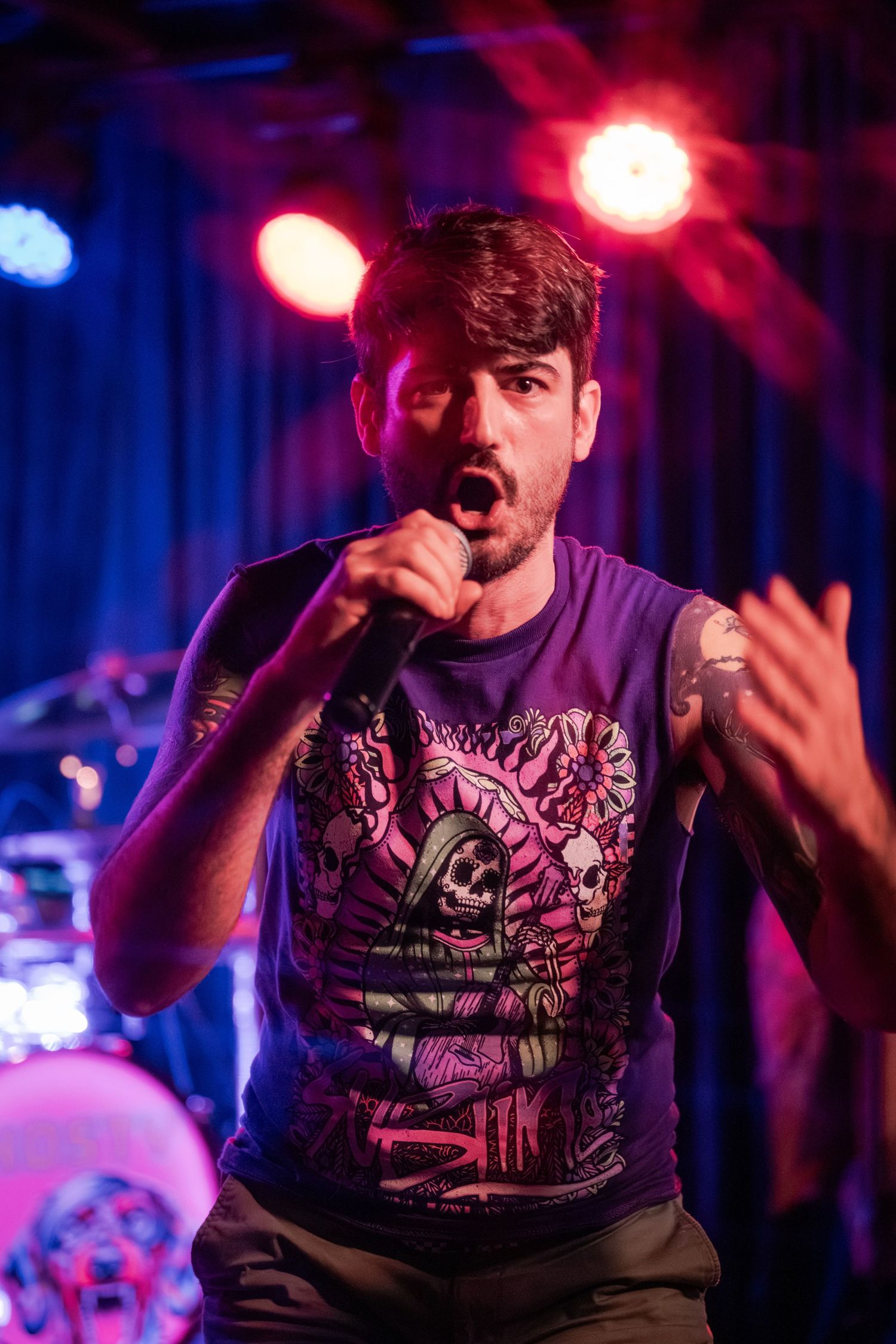 A performer sings passionately on stage wearing a purple graphic t-shirt under colorful red and blue stage lighting.