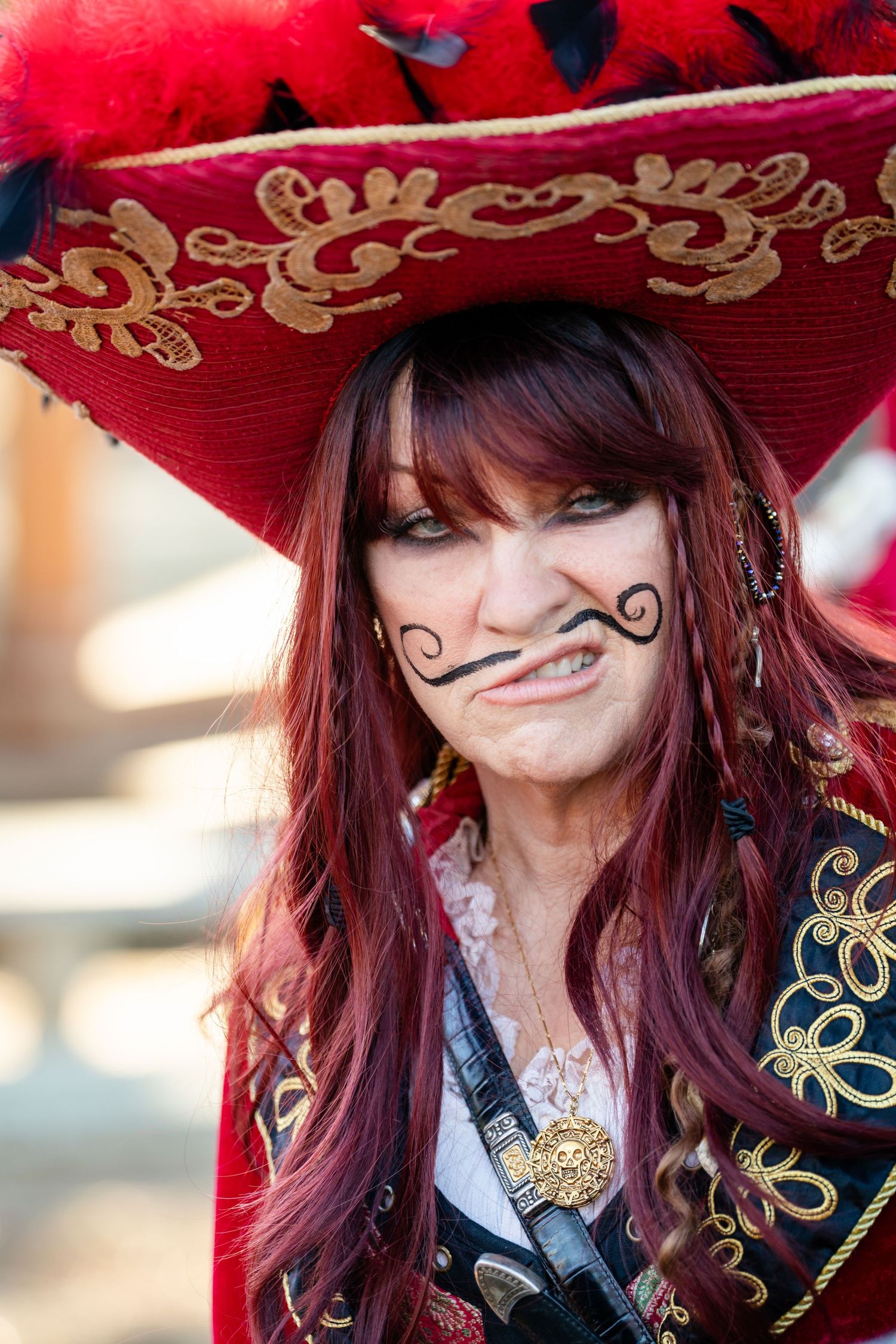 Person wearing a red and gold embroidered mariachi hat and white shirt smirks playfully at the camera.