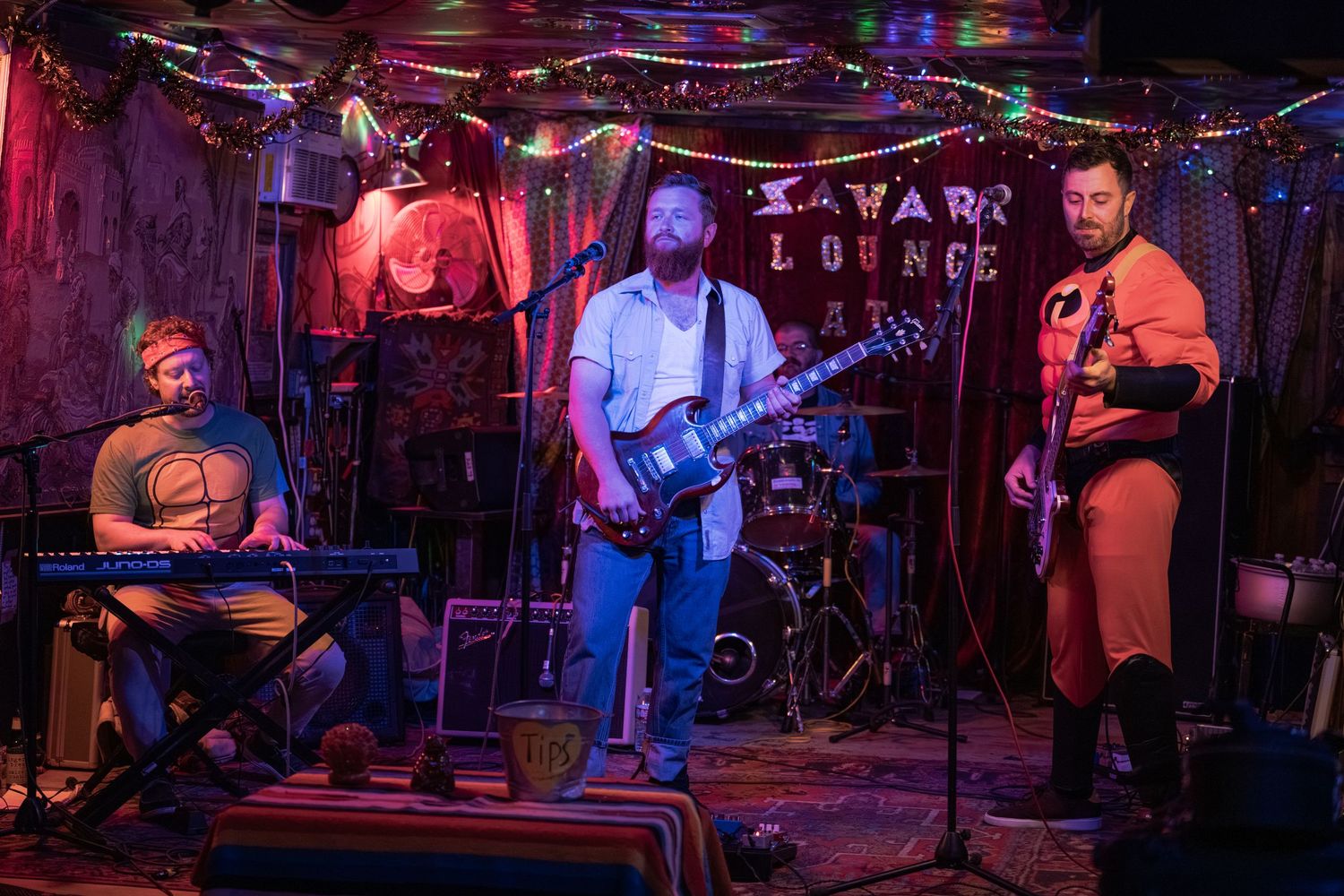 A band performs on stage at a dimly lit venue with red and blue lighting under a Kavana's Lounge sign.